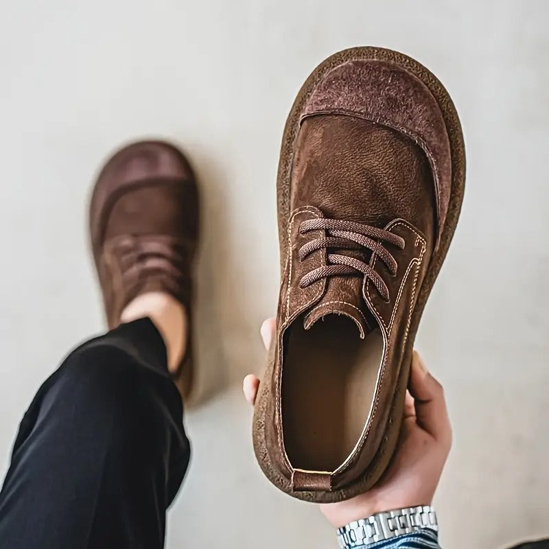 Dunkelbrauner Schuh in der Hand, von oben mit Blick auf Innenraum und Schnürung; der andere Schuh am Fuß unscharf im Hintergrund. 7