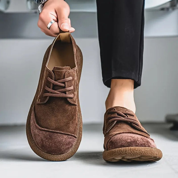 Dunkelbraune Schnürschuhe, ein Schuh wird an der Lasche gehalten, der andere wird zur schwarzen Hose getragen. 6