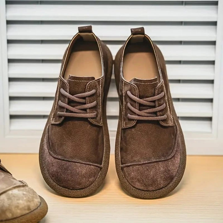 Paar dunkelbraune Schnürschuhe von vorn auf einem Holzbrett vor einer Lamellenwand, links ragt ein hellerer Schuh ins Bild. 6