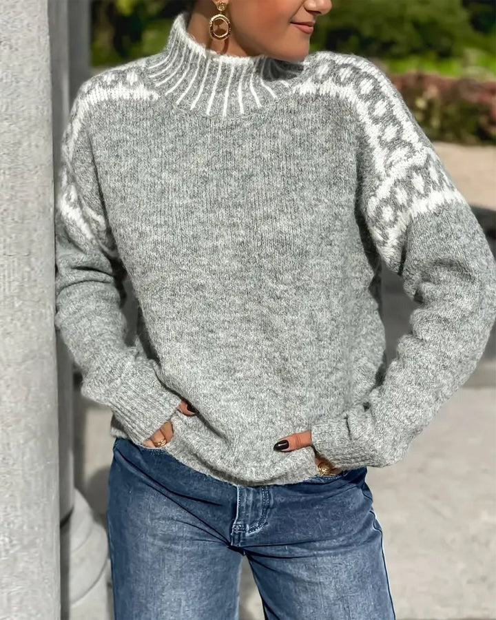 Grauer Strickpullover mit Stehkragen und weißem Zierstreifen über den Schultern, getragen zur Jeans vor einer Säule. 5