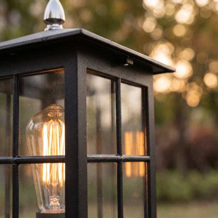 Nahaufnahme der Laterne mit leuchtender Glühfadenlampe, Glasscheiben, strukturiertem schwarzem Rahmen und glänzender Dachspitze vor weichem Hintergrund. 5