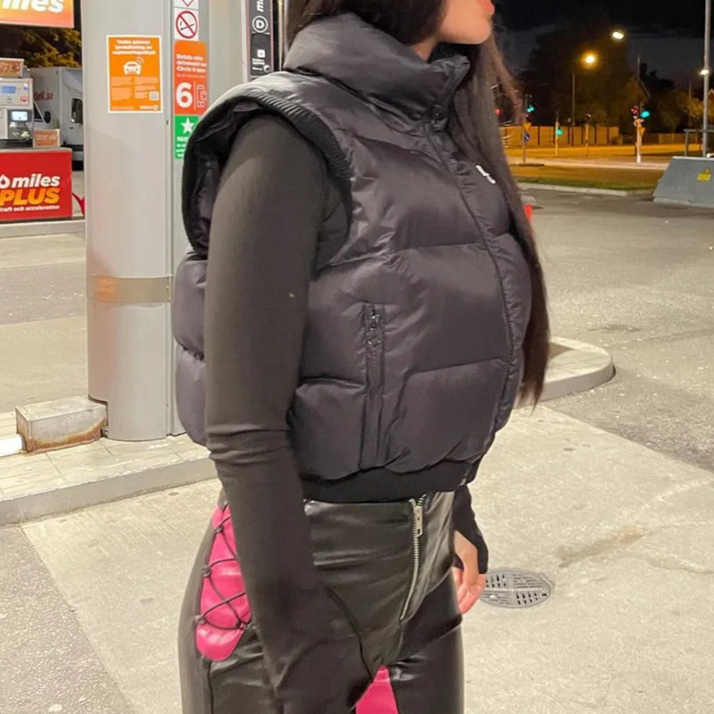 Frau in schwarzer Daunenweste und Lederhose an Tankstelle bei Nacht.
