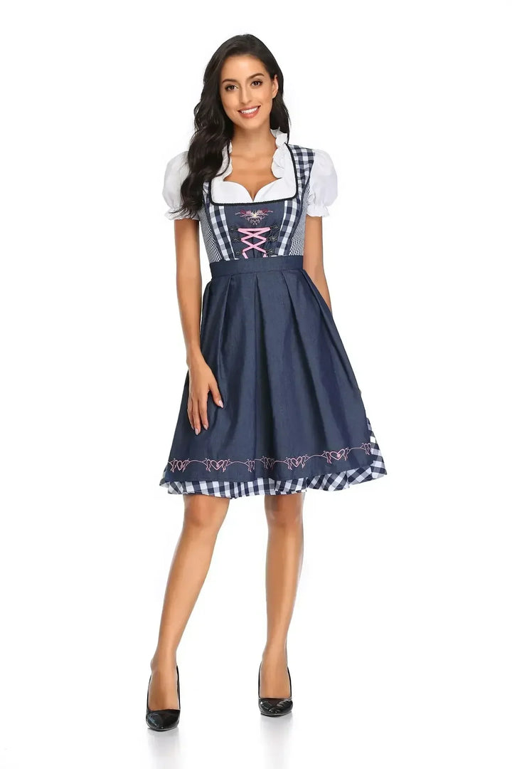 Frontansicht des Dirndls in Marineblau mit blau-weiß kariertem Oberteil, rosa Schnürung und bestickter Schürze, getragen von einer lächelnden Frau. 3