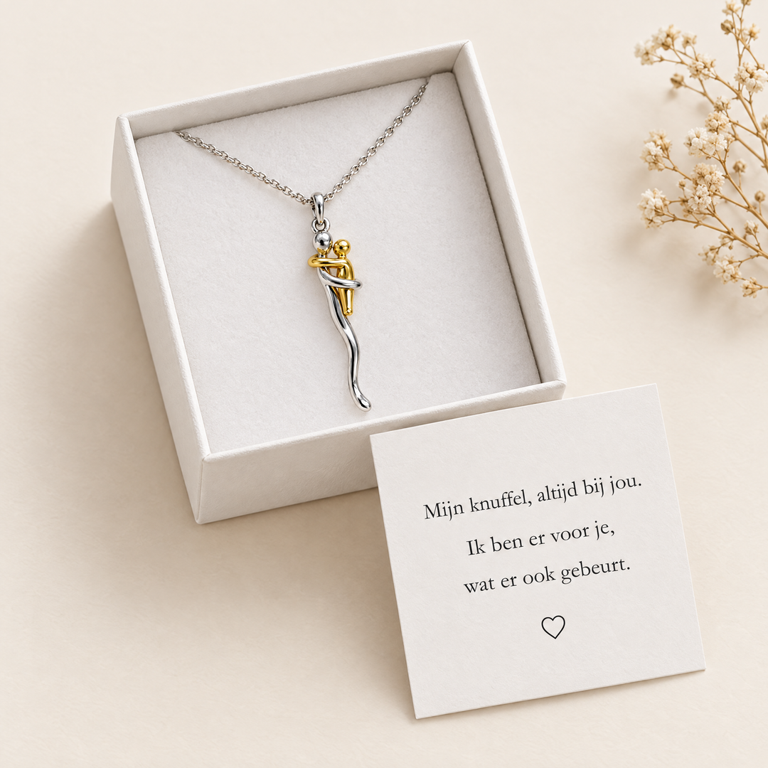 Halskette mit Umarmungs-Anhänger in weißer Geschenkschachtel auf Samteinlage, daneben eine Karte mit Botschaft und getrocknete Blüten. 2