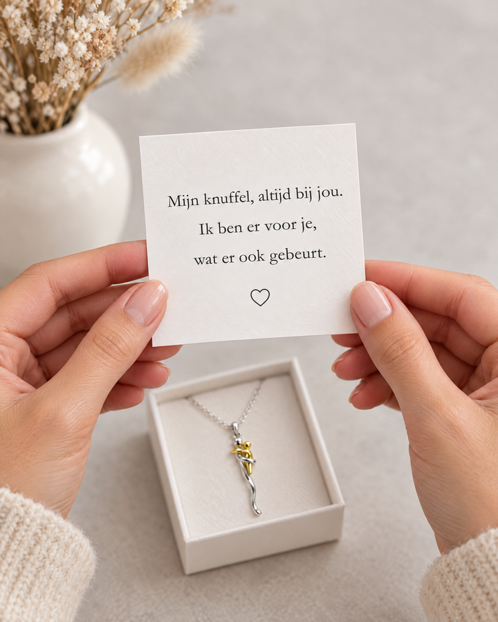 Hände halten eine Grußkarte über der geöffneten Geschenkschachtel, in der die Halskette mit Anhänger liegt, links eine Vase mit Trockenblumen. 6
