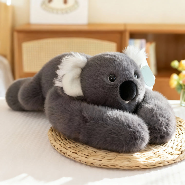 Graues Koala-Kuscheltier mit weißen, flauschigen Ohren liegt auf einer Strohmatte, die langen Arme umschlingen den Körper. 16