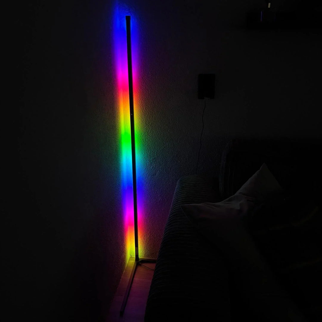 Leuchtende Eckleuchte im dunklen Raum mit Regenbogen-Farbverlauf an der Wand, rechts ein Sofa mit Kissen. 6
