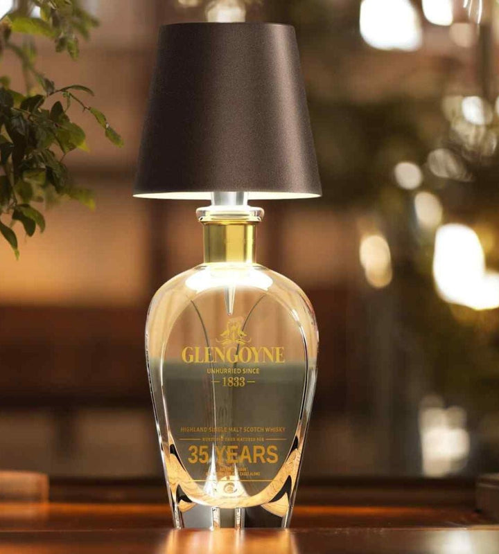 Dunkelbrauner Lampenschirm auf einer eckigen Whiskykaraffe aus Glas, die leuchtend auf einem Holztisch steht. 5