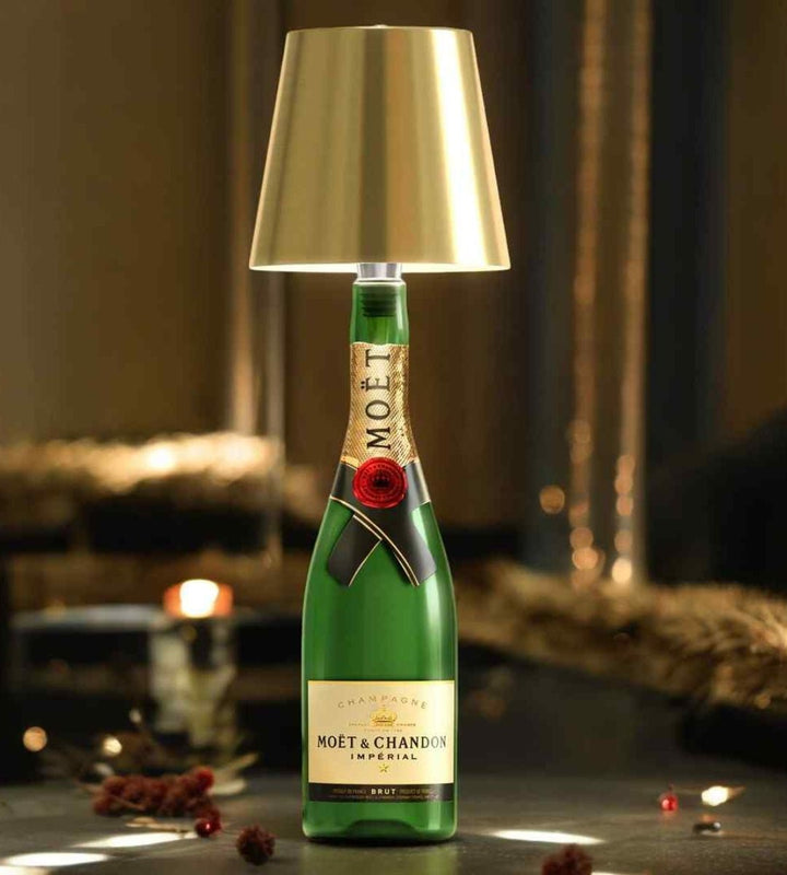 Goldener Lampenschirm auf einer grünen Sektflasche, die auf einem dunklen Tisch mit festlicher Weihnachtsdekoration und Lichterkette steht. 1
