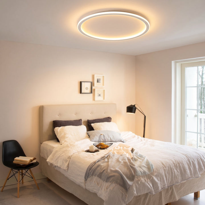 Leuchtender Deckenring über einem Schlafzimmer mit gepolstertem Bett, heller Bettwäsche, Stehlampe und Balkontür. 9