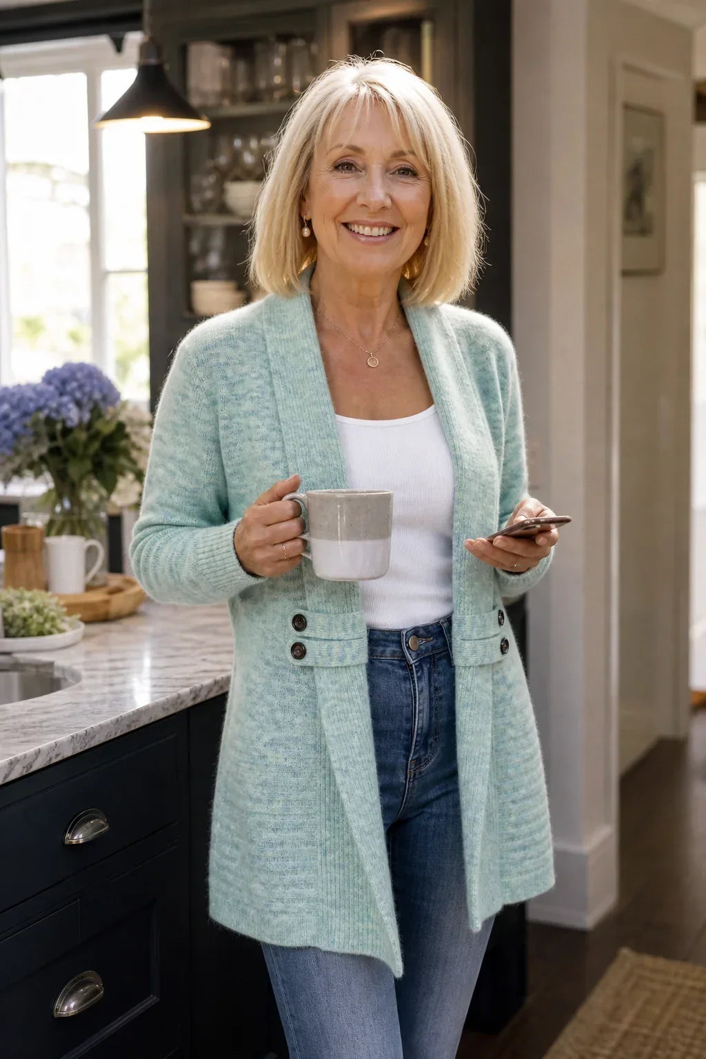 Lächelnde Frau in mintgrünem Cardigan hält in der Küche eine Tasse und ein Smartphone 4
