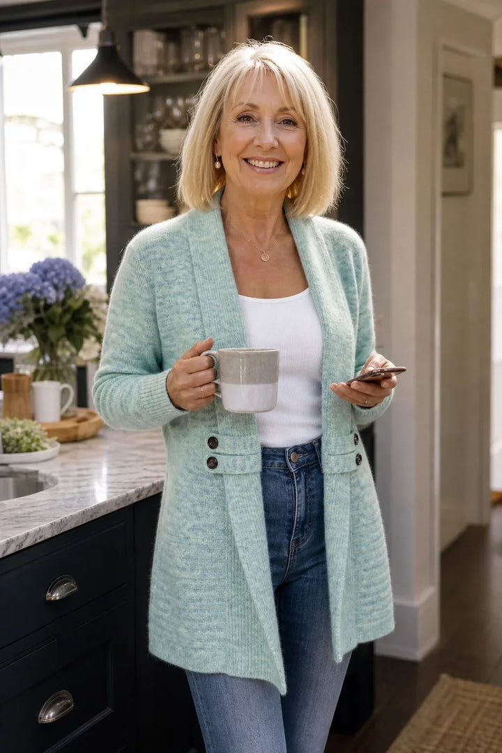 Lächelnde Frau in mintgrünem Cardigan hält in der Küche eine Tasse und ein Smartphone 4