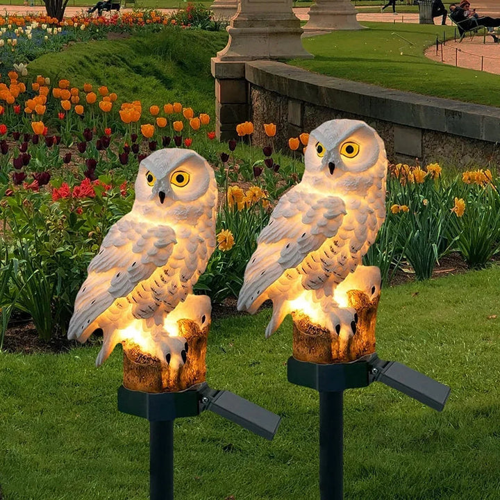 Zwei leuchtende weiße Eulen auf Erdspießen im Gras vor einem Blumenbeet mit orangen und gelben Tulpen und Narzissen. 1