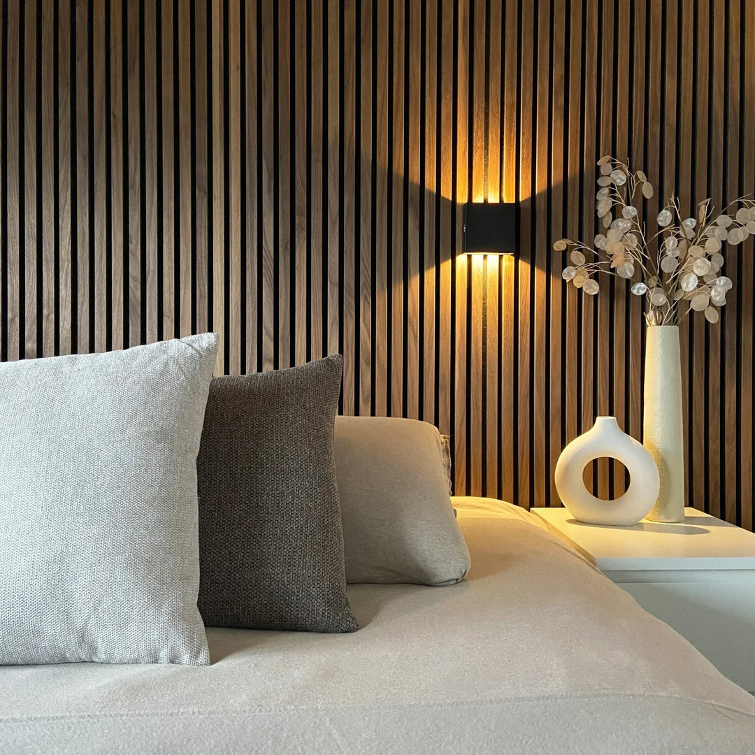 Schwarze Wandleuchte an einer Holzlamellenwand über dem Kopfteil eines Betts, daneben Kissen, weiße Vasen und getrocknete Zweige. 5