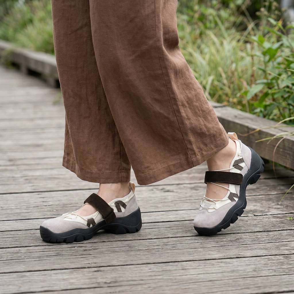 Beige Slipper mit dunkelbraunem Klettriemen, getragen mit weiter brauner Leinenhose auf einem Holzsteg neben Gräsern im Freien. 11