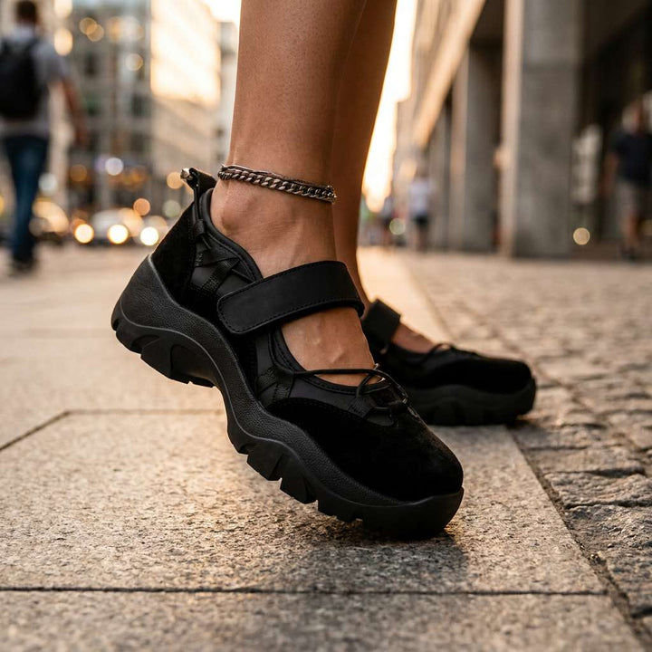 Schwarze Slipper mit Klettriemen und dicker Sohle, getragen mit Fußkettchen auf einem Steinpflaster in der Stadt bei Abendlicht. 13