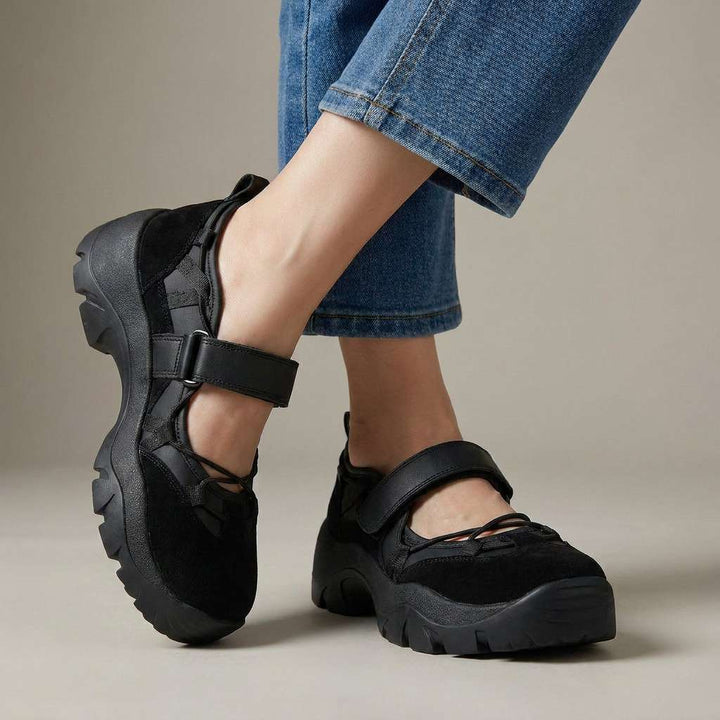 Komplett schwarze Slipper aus Veloursleder mit Klettriemen und kräftiger Profilsohle, getragen mit heller Jeans vor neutralem Studiohintergrund. 5