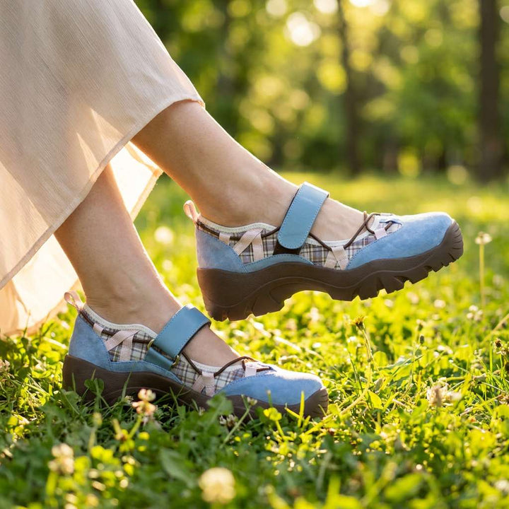 Hellblaue Slipper mit Klettriemen und kariertem Einsatz, getragen zu einem fließenden cremefarbenen Kleid auf einer sonnigen Blumenwiese. 8
