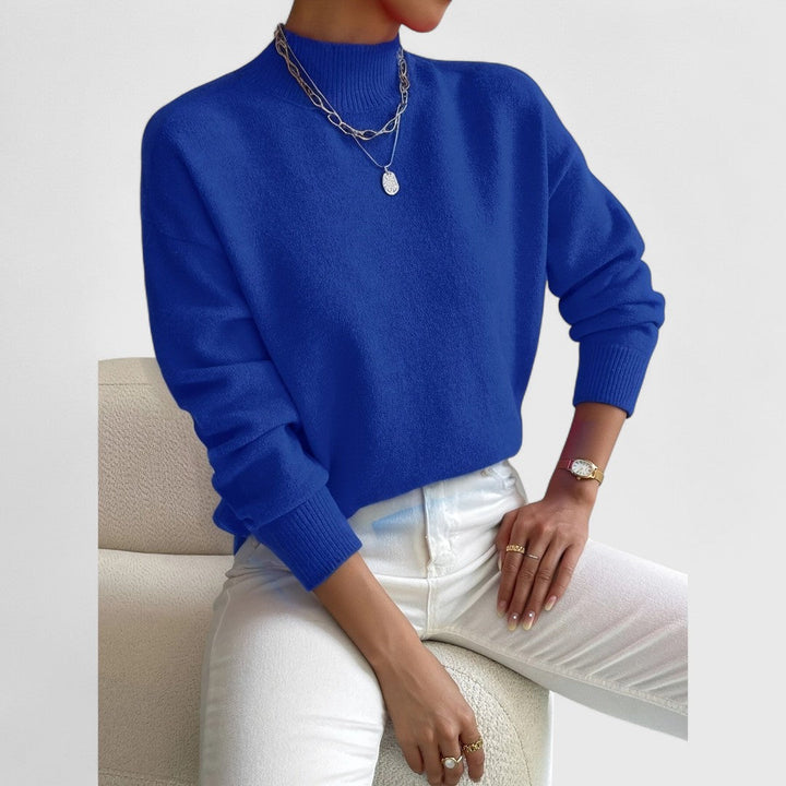 Königsblauer Pullover mit Stehkragen am Model, getragen mit weißer Hose, silbernen Ketten und goldener Armbanduhr. 11