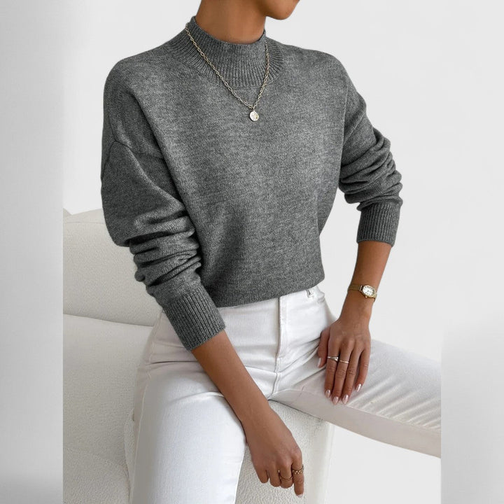 Grau melierter Pullover mit Stehkragen und Rippbündchen am Model, kombiniert mit weißer Hose, dünner Kette und Armbanduhr, leicht seitlich fotografiert. 12