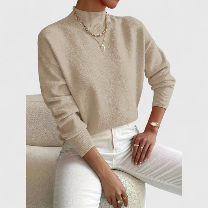 Beigefarbener Pullover mit Stehkragen am Model, kombiniert mit weißer Hose, goldenen Ketten und Ringen auf einem cremefarbenen Sessel. 7