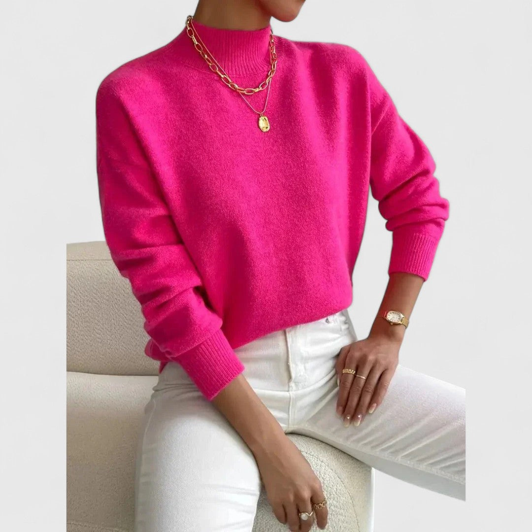Pinkfarbener Pullover mit Stehkragen und Rippbündchen am Model, kombiniert mit weißer Hose und goldenem Schmuck. 9