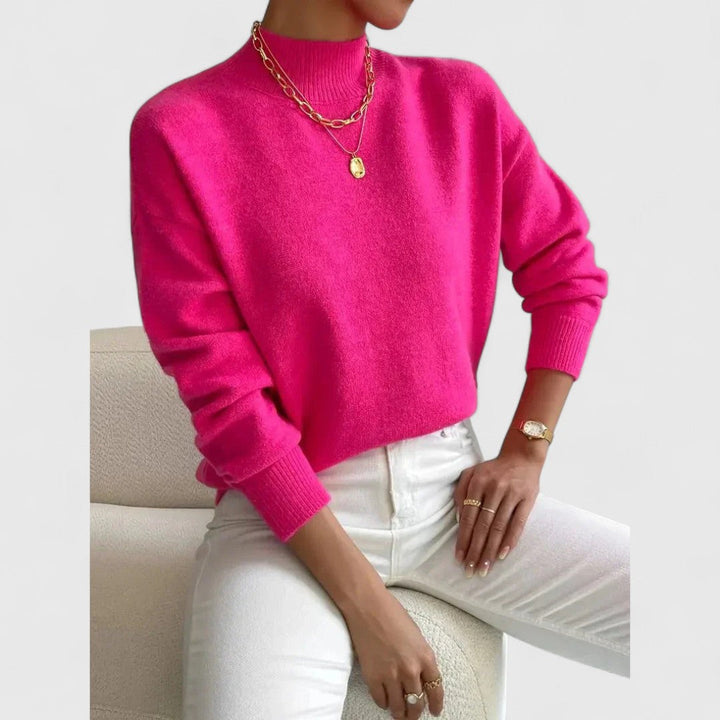 Pinkfarbener Pullover mit Stehkragen und Rippbündchen am Model, kombiniert mit weißer Hose und goldenem Schmuck. 9