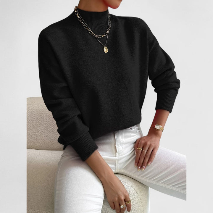 Schwarzer Pullover mit Stehkragen am Model, getragen mit weißer Hose, goldenen Ketten und Armbanduhr, sitzend auf hellem Sessel. 8