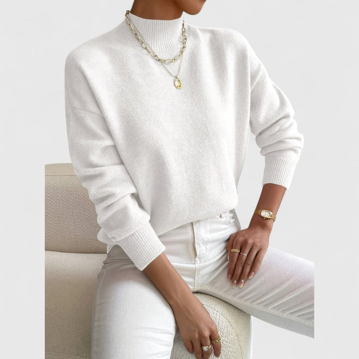 Weißer Pullover mit Stehkragen und Rippbündchen am sitzenden Model, kombiniert mit weißer Hose und goldenem Schmuck. 4