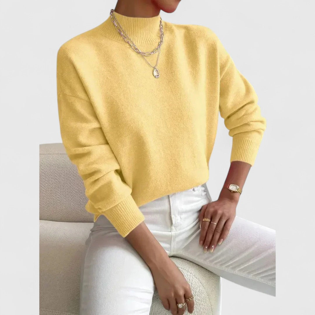Gelber Pullover mit Stehkragen und gerippten Bündchen, getragen vom sitzenden Model mit weißer Hose und Halsketten. 6