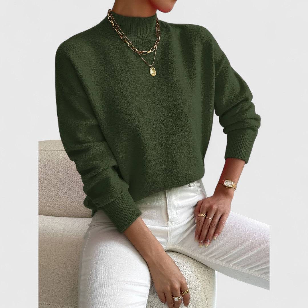Dunkelgrüner Pullover mit Stehkragen am sitzenden Model, kombiniert mit weißer Hose, goldenen Halsketten und Ringen. 10