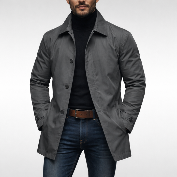 Frontansicht des grauen Mantels am Model mit schwarzem Rollkragen, brauner Gürtelschnalle und dunkler Jeans, Hände in den Manteltaschen. 2