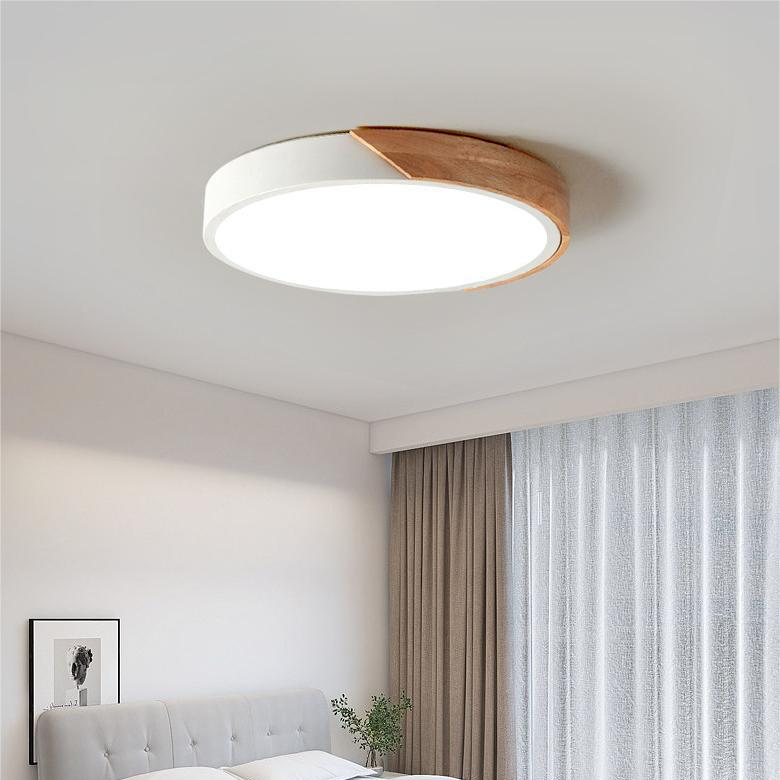 Weiße runde Deckenleuchte mit Holzsegment über einem Schlafzimmerbett mit grauem Kopfteil, Vorhängen und kleiner Zimmerpflanze. 3