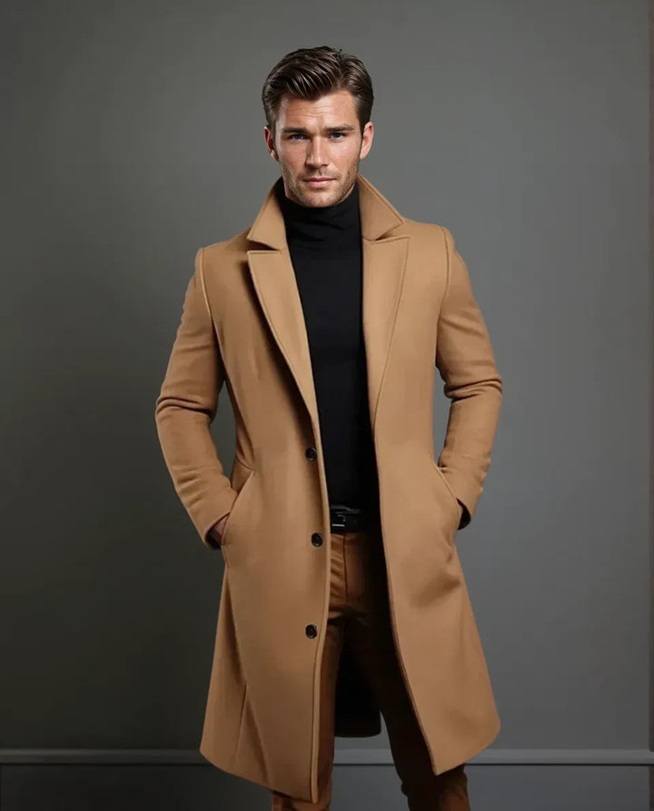 Ganzkörperansicht eines Models im kamelfarbenen langen Mantel mit schwarzem Rollkragenpullover und brauner Hose, Hände in den Manteltaschen. 5