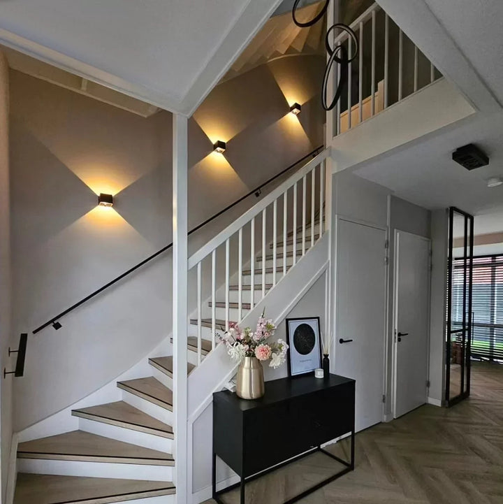 Drei schwarze Wandleuchten entlang einer Treppe mit weißem Geländer, im Flur steht eine schwarze Kommode mit Blumenvase. 7