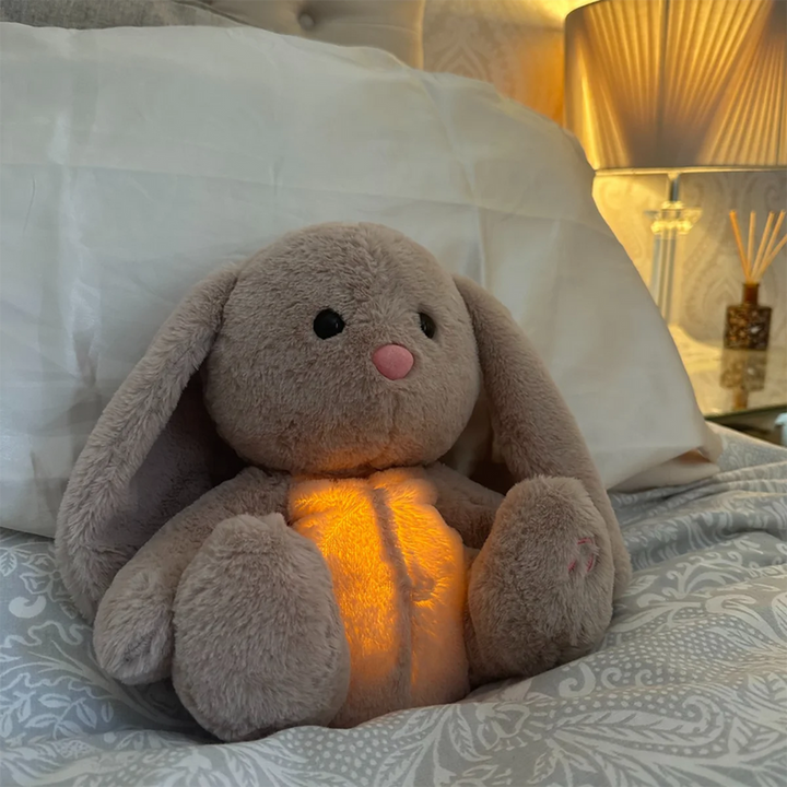 Hasen-Plüschtier mit orange leuchtendem Bauch lehnt auf einem Bett an einem weißen Kissen, im Hintergrund eine Lampe. 15