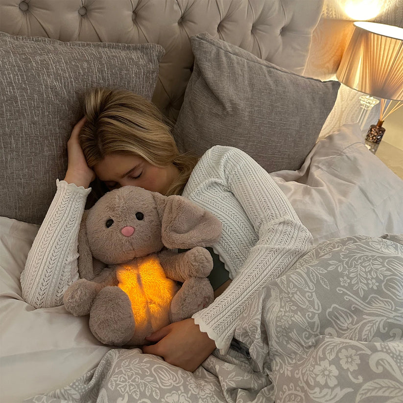 Schlafende Frau mit blonden Haaren im Bett, den Kopf an das Hasen-Plüschtier mit leuchtendem Bauch gelehnt. 16