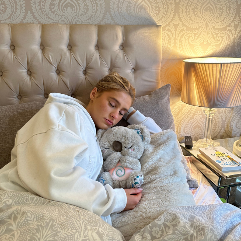 Junge Frau im weißen Hoodie schläft im Bett und hält das Koala-Plüschtier mit leuchtender Spirale am Bauch im Arm. 14