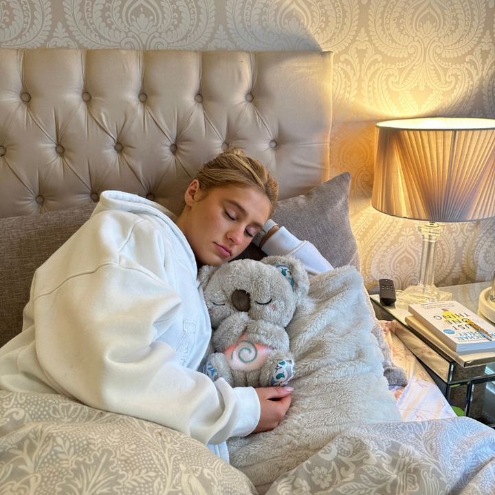 Junge Frau im weißen Hoodie schläft im Bett und hält das Koala-Plüschtier mit leuchtender Spirale am Bauch im Arm. 14