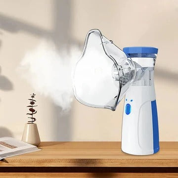 Weiß-blaues Handgerät mit angesetzter durchsichtiger Maske auf einem Holztisch, aus der feiner Nebel austritt, daneben Buch und kleine Pflanze. 1
