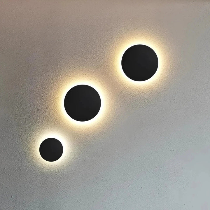 Drei schwarze, runde Wandleuchten in verschiedenen Größen strahlen warmes Licht als leuchtenden Ring an eine helle, strukturierte Wand. 2