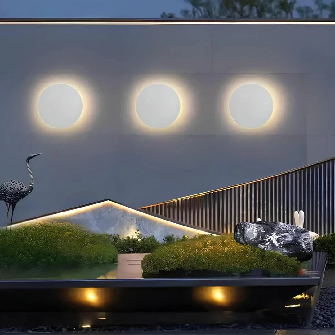 Drei gleich große weiße Rundleuchten nebeneinander an einer Gartenmauer in der Dämmerung, davor ein beleuchtetes Gartenarrangement mit Moos und Steinen. 5