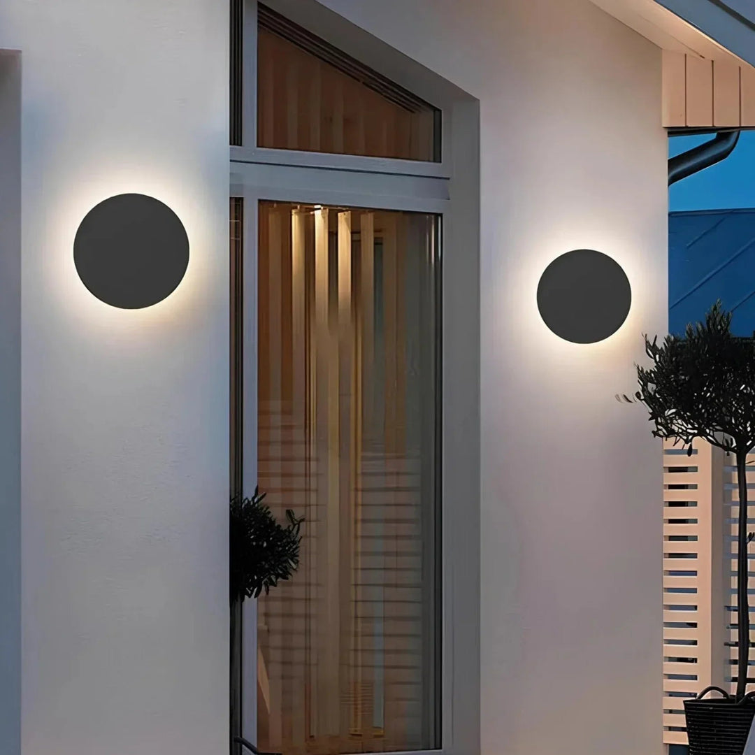 Zwei runde schwarze Wandleuchten an einer weißen Hausfassade neben einer Glastür, abends mit warmem Lichtkranz. 3