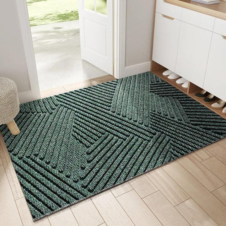 Dunkelgrüne Fußmatte mit erhabenem geometrischem Streifenmuster im Eingangsbereich, neben Schuhschrank und offener Tür auf Holzboden. 1