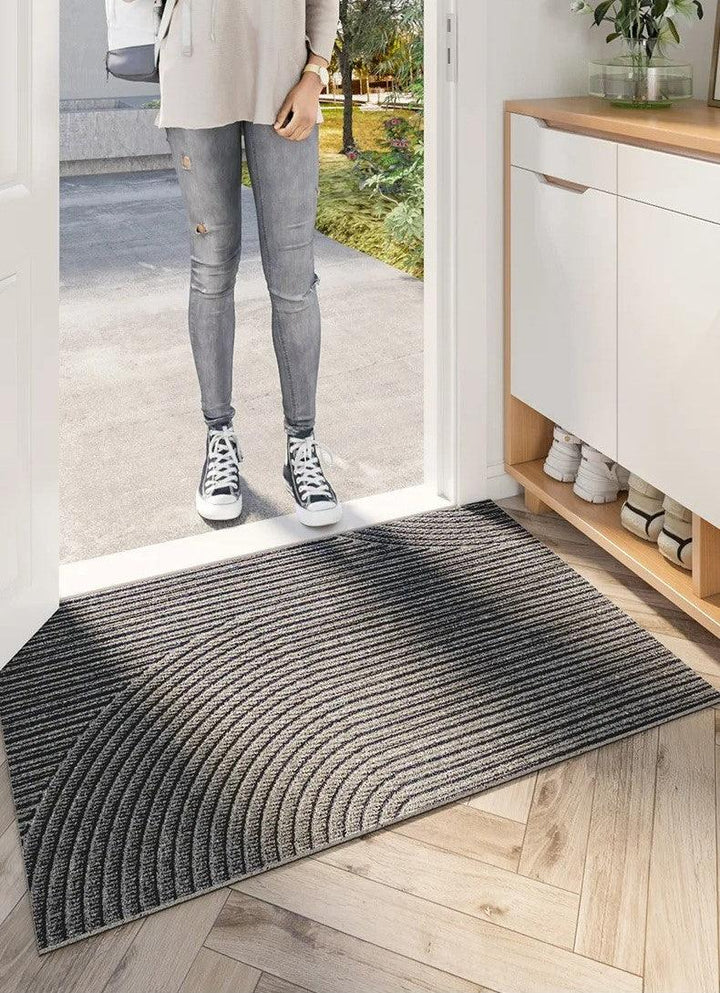 Graue Fußmatte mit Bogenmuster im Türrahmen, davor steht eine Person in Jeans und Sneakern vor dem Eingang. 5