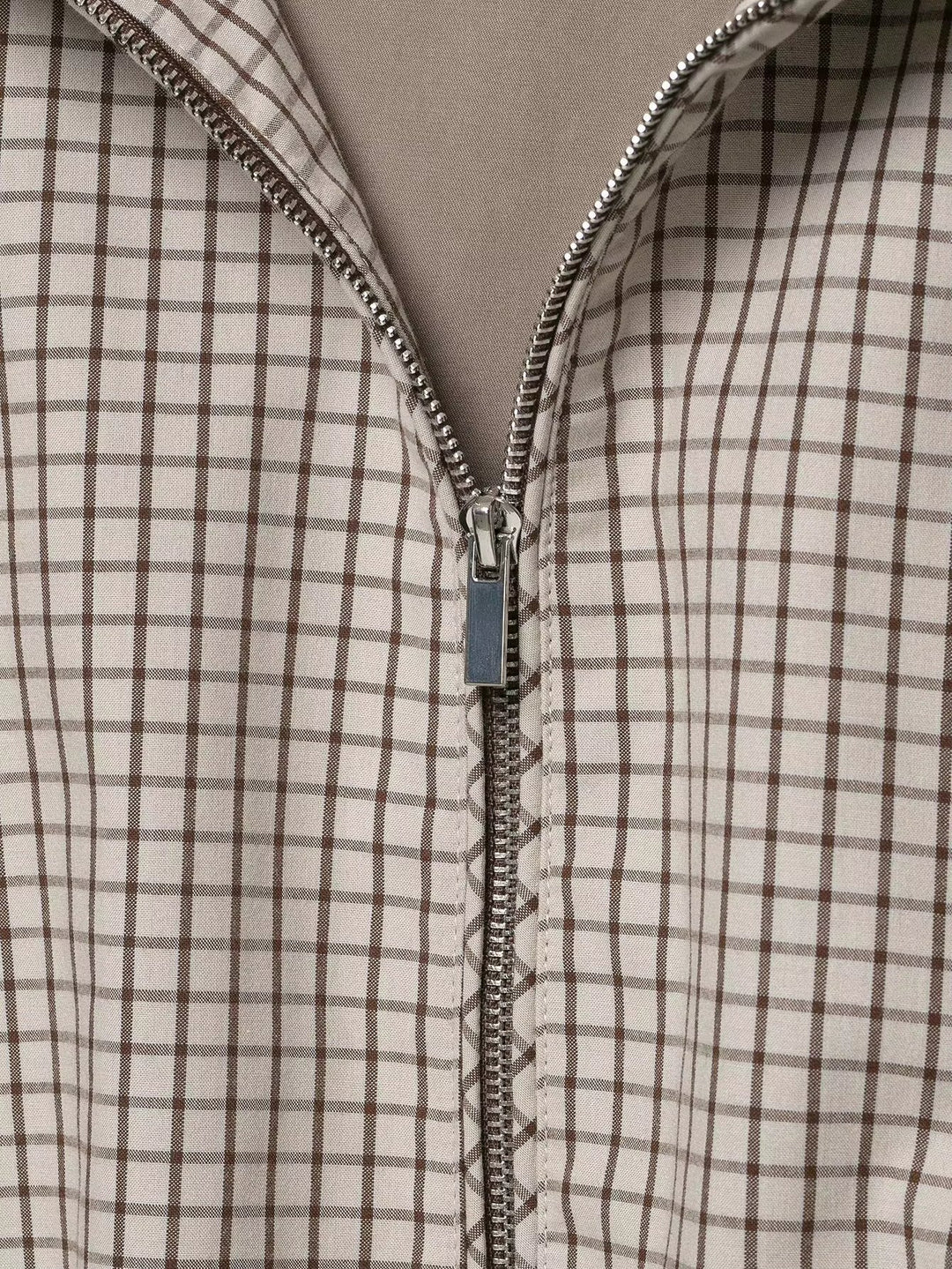 Detailaufnahme des halb geöffneten silbernen Reißverschlusses vor dem feinen braun-beigen Karomuster, darunter das taupefarbene Futter. 6