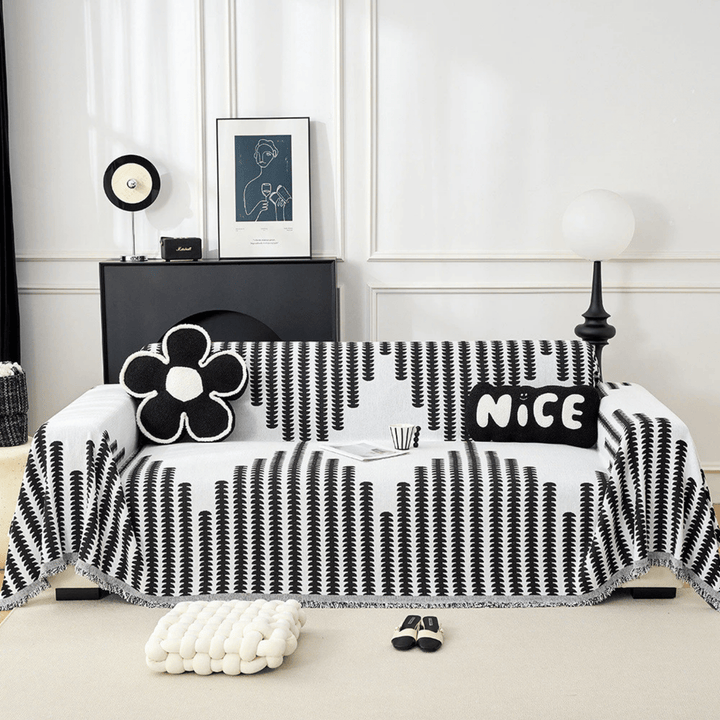 Schwarz-weiß gemusterter Sofabezug mit Fransenkante, frontal auf einem Sofa im hellen Wohnzimmer, dekoriert mit schwarzen Zierkissen. 1