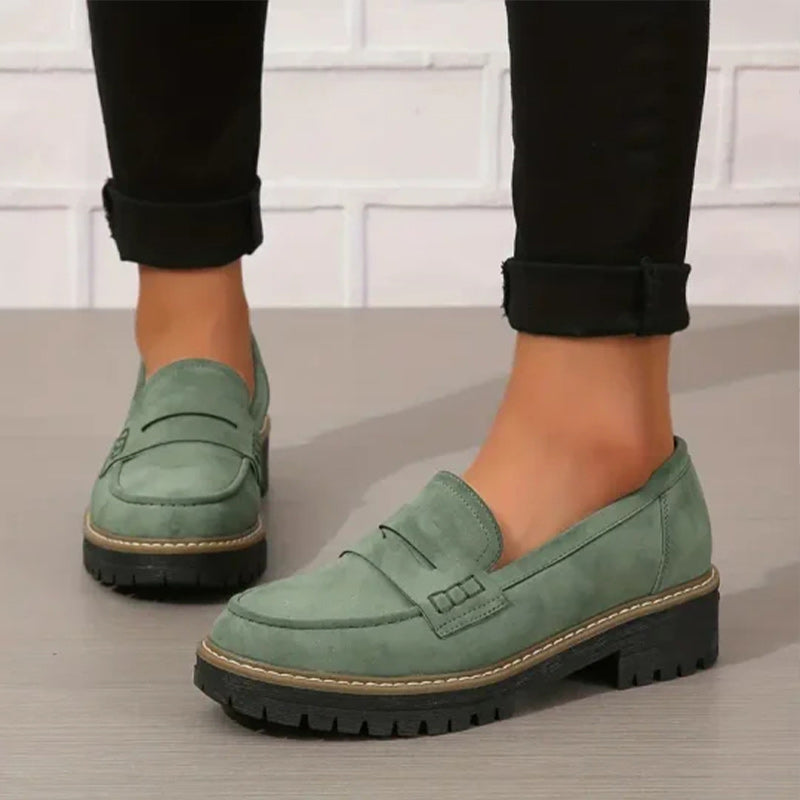 Grüne Wildleder-Loafer mit schwarzer Profilsohle, getragen zu schwarzer Hose mit umgeschlagenem Saum vor weißer Backsteinwand. 1