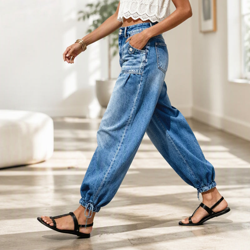Seitenansicht einer gehenden Frau in blauer Barrel-Jeans mit Handtasche, Saumkordeln und schwarzen Riemensandalen in einem hellen Raum. 7