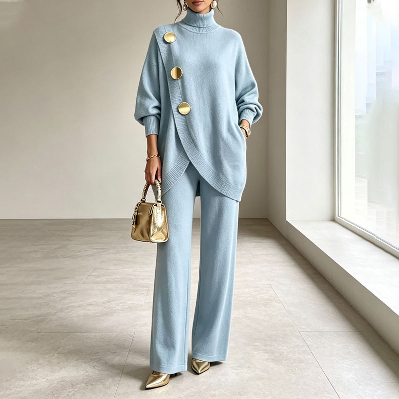 Model in hellblauem Strickset mit Rollkragen, goldenen Knöpfen und weiter Hose, goldene Handtasche in der Hand, vor einem Fenster. 1